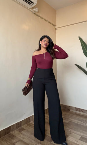 Woman in burgundy top and premium black sculpted trouser standing indoors against a plain wall.